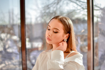 A young girl with makeup dressed in a white shirt sits near a large panoramic window.