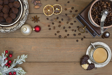 Vintage manual coffee mill, coffee beans, retro metal cup with coffee, silver plate with chocolate truffles and Christmas decorations with snowy pine branches and dry spices on a brown wooden planks.