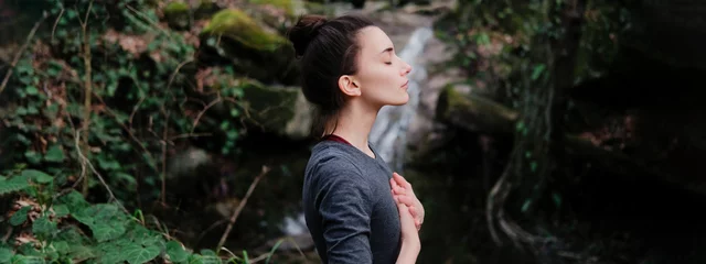 Fotobehang Young woman practicing breathing yoga pranayama outdoors in moss forest on background of waterfall. Unity with nature concept. © yolya_ilyasova