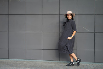 Fototapeta na wymiar Spring modern collection of spring women's clothing. Pretty young serious woman with long hair wearing youth grey oversize dress and elegant hat in windy day. Shot on urban grey wall background
