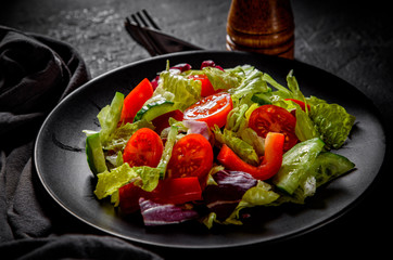 various fresh mix salad leaves with tomato, cucumber and red pepper on a black plate on Dark grey black slate background