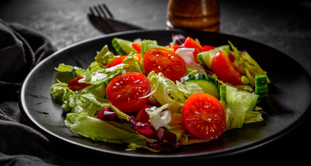 various fresh mix salad leaves with tomato, cucumber and red pepper on a black plate on Dark grey...