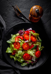 various fresh mix salad leaves with tomato, cucumber and red pepper on a black plate on Dark grey black slate background