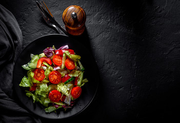 various fresh mix salad leaves with tomato, cucumber and red pepper on a black plate on Dark grey black slate background