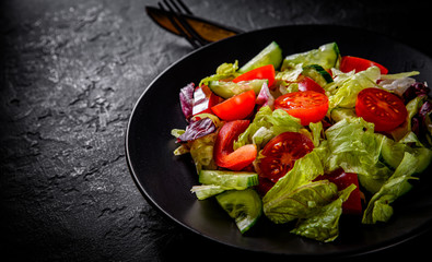 various fresh mix salad leaves with tomato, cucumber and red pepper on a black plate on Dark grey black slate background