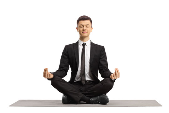 Young man in a black suit with closed eyes sitting with crossed legs and making a meditation pose on a mat - Powered by Adobe