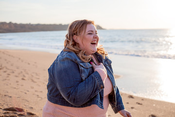 Beautiful overweight woman walking on the sandy beach. Plus size girl enjoy warmth sunset with romantic mood. Fat model dressed jeans jacket and pink knitted sweater - obrazy, fototapety, plakaty