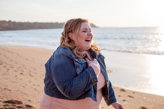 Beautiful overweight woman walking on the sandy beach. Plus size girl enjoy warmth sunset with romantic mood. Fat model dressed jeans jacket and pink knitted sweater