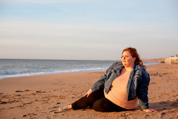 Fototapeta na wymiar Back view of beautiful overweight woman sitting on the sandy beach. Plus size girl enjoy warmth sunset with romantic mood. Fat model dressed jeans jacket and pink knitted sweater
