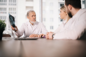 Head doctor of the hospital department explains to his colleagues result of patient's MRI scan examination. Elderly doctor with white beard holds X-ray in his hands. Concept of team medical work.