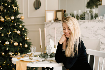 Christmas portrait of a smiling blonde girl in a black dress on the background of Christmas decor at a table in an elegant interior. A woman is preparing to celebrate Christmas and New year