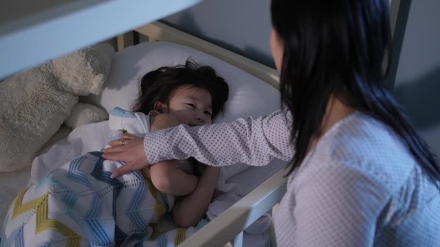 Close-up of caring chinese mom stroking and kissing asleeping little daughter and wishing her good night while sitting next to her bed. Preadolescent girl hugging beloved mother while going to bed