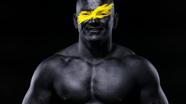 Man bodybuilder athlete with yellow color on face art and black body paint plays muscles on the chest. Colorful portrait of the guy with bodyart.