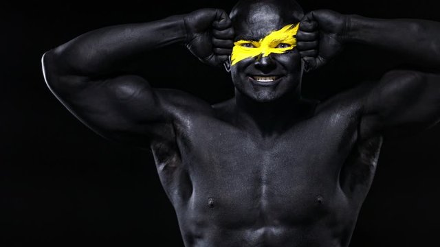 Man bodybuilder athlete with yellow color on face art and black body paint plays biceps muscles. Colorful portrait of the guy with bodyart.
