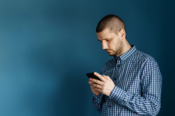 Caucasian man wearing shirt standing in front of the blue background wall using smart phone mobile to send messages sms texting or browsing internet front view