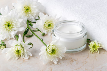 Fototapeta na wymiar Facial cream in an open glass jar and white chrysanthemum flowers next to white terry towel. Spa, beauty, skincare and cosmetology concept.