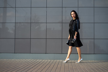 Obraz na płótnie Canvas Pretty beautiful young woman wearing elegant black dress and fashionable black fringed leather jacket holding stylish sunglasses in the hand. Urban wall on background. Copy space.