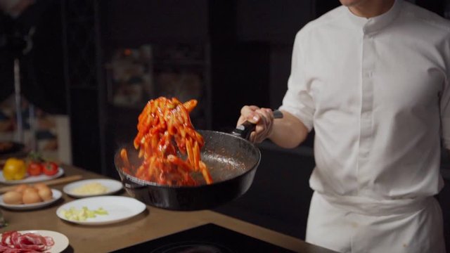 Panning tilt down medium shot of unrecognizable young chef tossing pasta with tomato sauce in pan in slow motion while cooking in restaurant kitchen