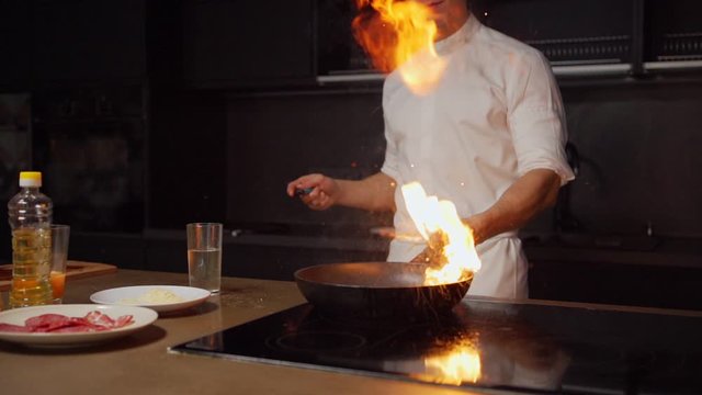 Slow motion medium shot of unrecognizable young chef using lighter to flambe meat in pan with big flame preparing dish on stove in restaurant kitchen