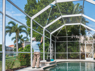 Handyman cleaning outdoor pool cage enclosure with pole brush. Screened swimming pool lanai maintenance and screen repair. - Powered by Adobe