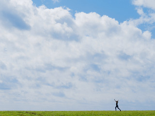 Fototapeta premium Sporty girl is full of joy and running on a green field with her hands up high with a blue sky with white clouds on the background. 