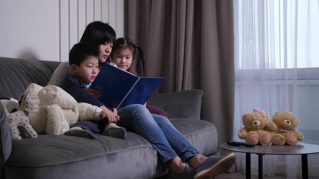 Cute children attentively listening to mom reading book during leisure in domestic room. Responsible chinese mother spending free time for development of own preadolescent daughter and son