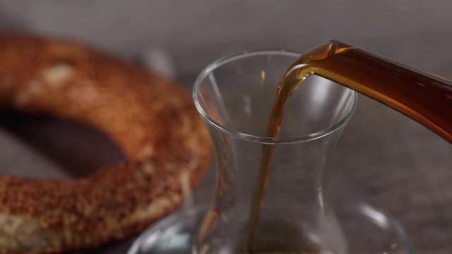 Pouring turkish tea into the cup with teapot and turkish simit is at background