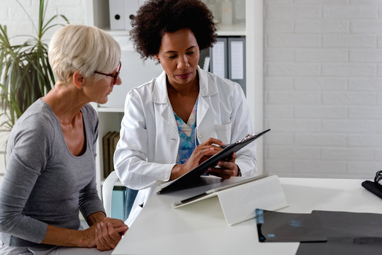 A Female Doctor Sits At Her Desk And Chats To An Elderly Female Patient While Looking At Her  Test Results