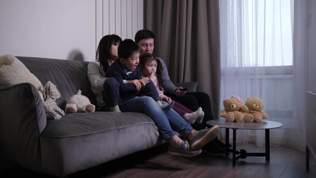 Happy chinese parents together with cute kids choosing TV program to watch while spending free time at home. Cheerful asian couple and their elementary age children watching TV during domestic leisure