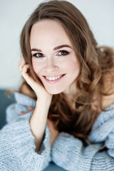 Close-up portrait of attractive young European smiling girl with light brown hair in light blue clothes. She poses sitting on a light blue chair. Studio shooting.  Light background.