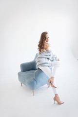 Attractive European girl with light brown hair in pastel clothes and beige shoes on a heel poses sitting on a light blue chair. Studio shooting.  Light background.