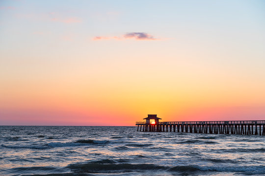 Naples, Florida vibrant red yellow sunset in gulf of Mexico with sun inside framing Pier wooden jetty on horizon and dark silhouette ocean waves wide angle view