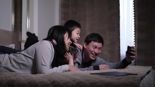 Closeup of cheerful asian parents with cute little kids posing for funny photo on cellphone while lying on bed. Happy dad, mom, little daughter and son posing for selfie photo on smartphone in bedroom