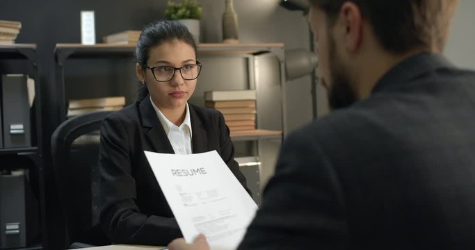 Back view of mature bearded man in formal clothing holding resume and talking about himself at job interview. Female office manager in eyeglasses listening carefully new applicant.