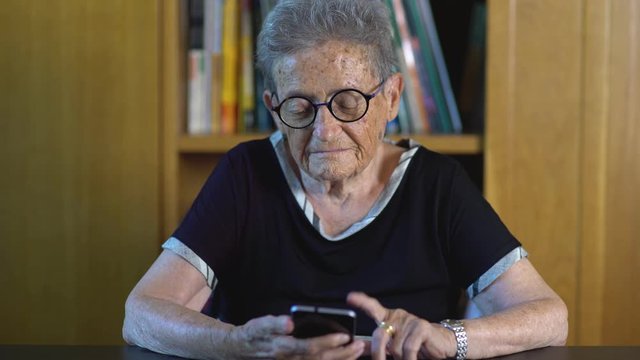 Nice old woman with round glasses using joyfully a smartphone at home living room near a bookcase. Technological ederly lady surfing the internet with a mobile phone. Cine lens.