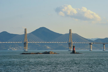 Obraz premium A bridge with two feature pylons stetches over a bay. Two rocky islands are in the foreground, one with a red channel marker. Mountains are in the background, with buildings on the foreshore. The sky 