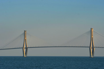 Two concrete pylons with radial supportive wires form a feature of a suspension bridge crossing a harbour. two tankers can be seen on the horizon. The sky is blue, with a haze near the horizon.