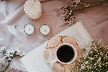A cup of coffee on a stand surrounded by flowers and candles of different sizes and shapes