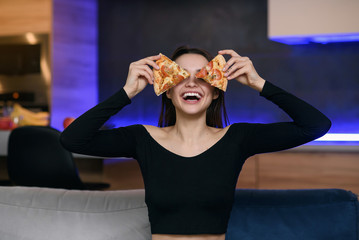 Excited happy young woman in kitchen plaing with pizza slices. Cover eyes with food. Carefree housekeeper very playful. Wear white dressing gown. Careless houseswife.