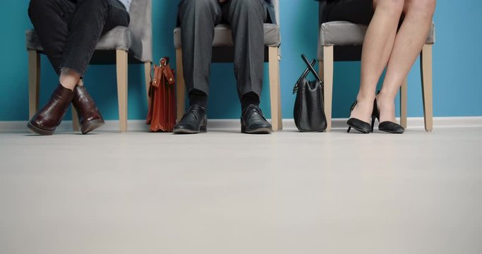 Close up of applicants legs in trendy shoes waiting for job interview at hall with blue wall and beige floor. Female and male competitors feeling stressed during expectation