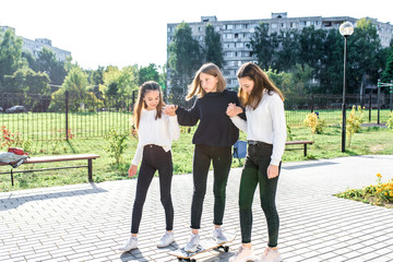 Three teenage girls, in summer in city, ride skateboard, rest break after school weekend. Casual warm clothing sweater jeans. Emotions of joy fun entertainment. Background lawn trees buildings.