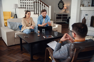 Close up of male psychologist sits at chair and listens depressed young couple at stylish cozy...