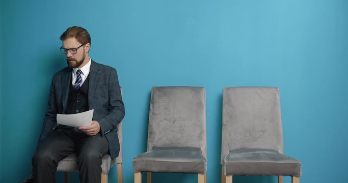 Serious mature man in trendy suit and eyeglasses preparing for job interview while waiting at blue hall. Bearded applicant reading his resume while sitting on grey chair