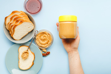 Creamy peanut paste in glass jar with yellow cap in female hand and peanuts in the peel scattered...