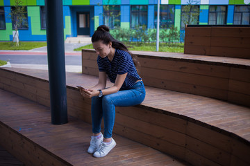 A brunette girl, student with long hair, with large wrist watch, in jeans, sits on wooden steps and checks a smartphone.