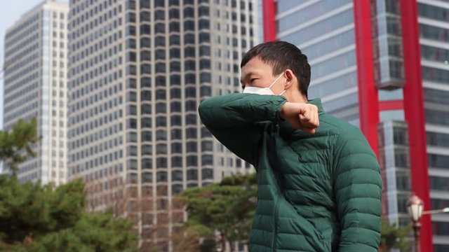 Asian man in white mask coughs and covers his face with his hand on the background of the city.