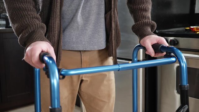 At home, a disabled older man walking with a walker slowly in his kitchen. Rollator walking frame concept.