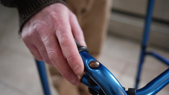 At home, a disabled older man with trembling hands walking with a walker slowly helped by the rollator walking frame. Parkinson disease, shaking hands concept.