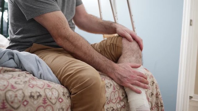 Man with an injury to his leg massaging it while sitting on his bed with crutches on the side.