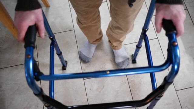 At home, a disabled older man walking with a walker slowly in his kitchen. Rollator walking frame concept.